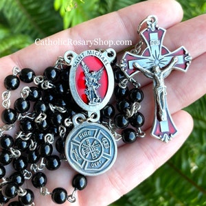 May include: Black rosary beads with a silver Saint Michael medal, a silver fire department medal, and a silver crucifix with a red and white enamel design. The crucifix has the inscription "INRI".