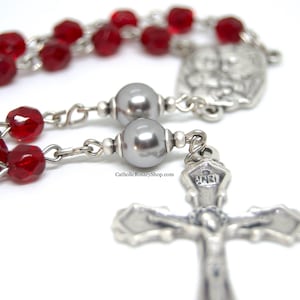 May include: A silver rosary with red glass beads and a silver crucifix. The crucifix has the inscription "INRI" on the crossbar.
