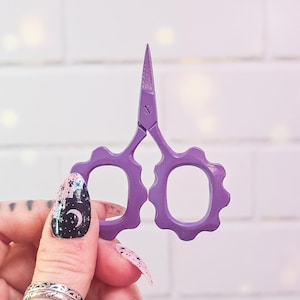 Puede incluir: Unas tijeras moradas para manualidades con un diseño decorativo de mango ondulado. Las tijeras se sostienen sobre un fondo de ladrillos blancos. Son pequeñas y afiladas, ideales para tareas de corte detalladas.