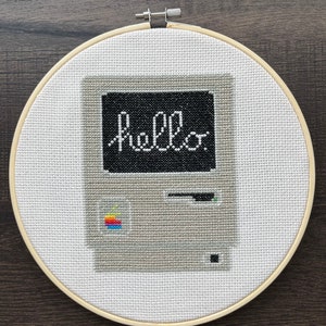 May include: Cross-stitch embroidery of a vintage Apple computer with a black screen displaying the word "Hello".