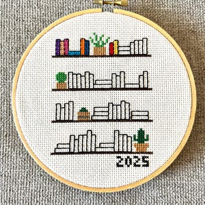 Könnte beinhalten: Ein weißer Kreuzstich-Stickrahmen mit einem Design aus vier Regalen, die mit Büchern und Topfpflanzen gefüllt sind. Das unterste Regal hat das Jahr 2025 aufgestickt.