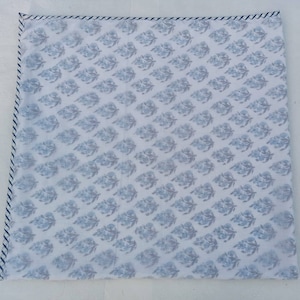 May include: Square white textile featuring a repeating blue floral design. The edges are finished with a blue and white striped border. The fabric has a soft, slightly translucent appearance.