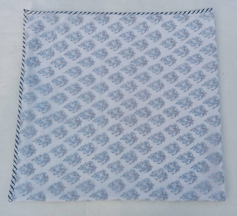 May include: Square white textile featuring a repeating blue floral design. The edges are finished with a blue and white striped border. The fabric has a soft, slightly translucent appearance.