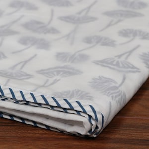 May include: White fabric with a repeating gray floral pattern. The edge of the fabric is trimmed with a blue and white striped border. The fabric is folded, showing the pattern and border.