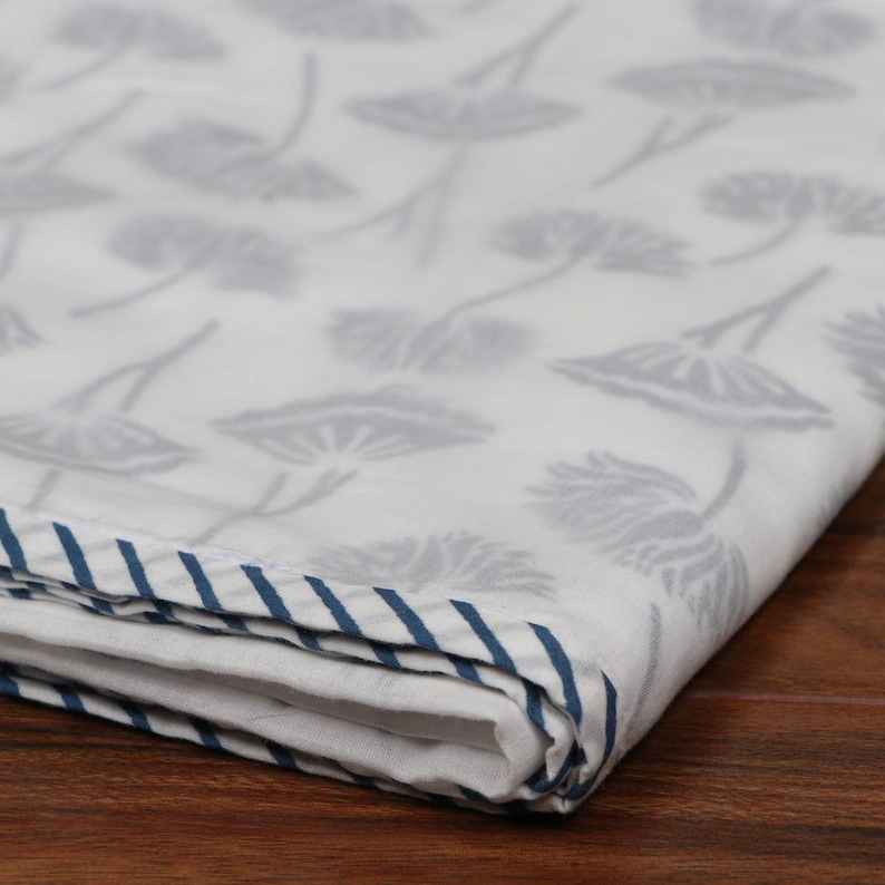 May include: White fabric with a repeating gray floral pattern. The edge of the fabric is trimmed with a blue and white striped border. The fabric is folded, showing the pattern and border.