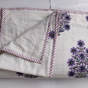 May include: White cotton quilt with a purple and green floral print. The quilt has a purple and white striped border.