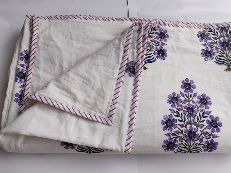 May include: White cotton quilt with a purple and green floral print. The quilt has a purple and white striped border.