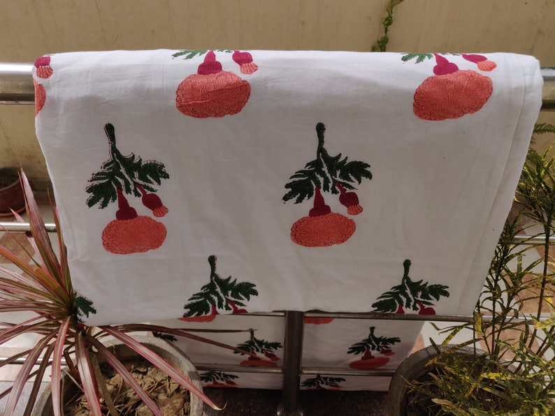May include: White cotton fabric with a repeating pattern of pink and green floral designs. The fabric is folded and draped over a metal bar.