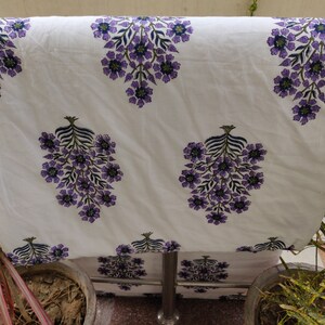 May include: White cotton fabric with a purple floral print. The fabric has a repeating pattern of purple flowers with green leaves.