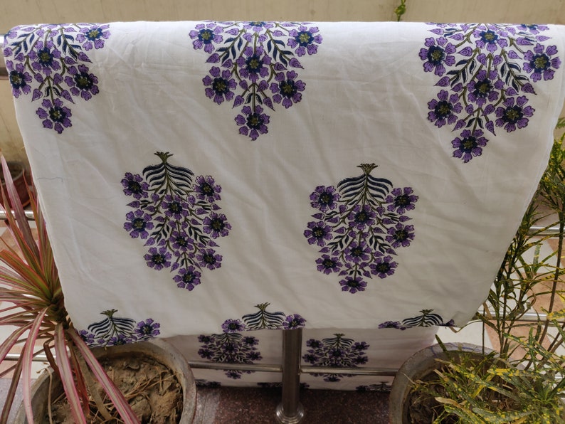 May include: White cotton fabric with a purple floral print. The fabric has a repeating pattern of purple flowers with green leaves.