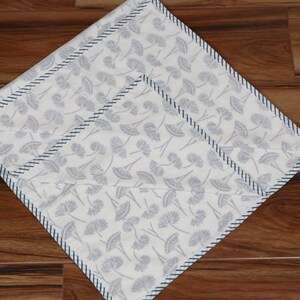 May include: Two white square cloth napkins with a gray floral pattern. Each napkin has a dark blue stitched border. The napkins are arranged diagonally on a wooden surface.