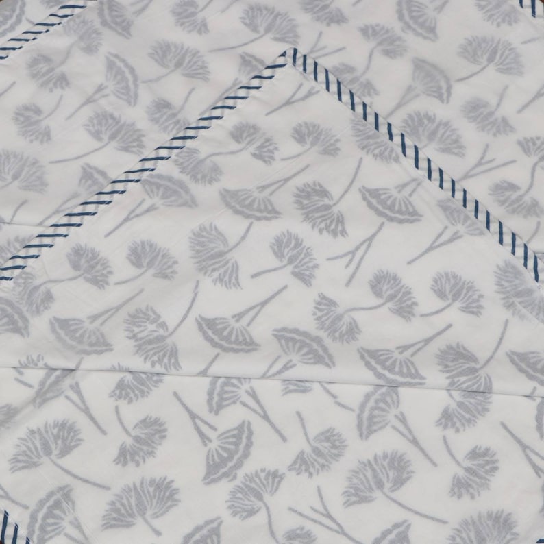 May include: White fabric with a repeating pattern of gray floral designs. The edges are trimmed with a navy blue stitched border. The fabric appears to be folded, creating a layered effect.