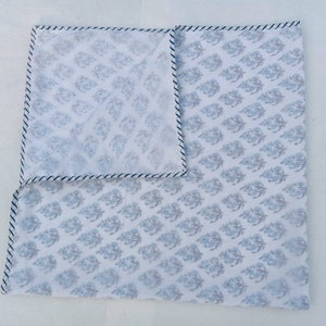 May include: White square textile featuring a repeating blue floral design. The edges are trimmed with a thin blue and white striped border. The textile is folded, showing the pattern on both sides.