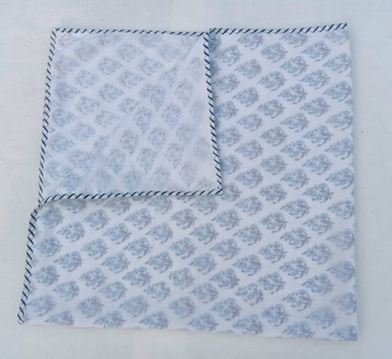 May include: White square textile featuring a repeating blue floral design. The edges are trimmed with a thin blue and white striped border. The textile is folded, showing the pattern on both sides.