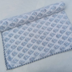 May include: Rolled white fabric featuring a repeating blue floral design. The edge is finished with a navy and white striped border. The material appears to be cotton or a similar fabric.