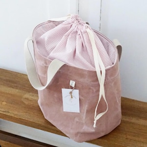 Könnte beinhalten: Eine rosa Canvas-Tragetasche mit weißen Henkeln und einer rosa-weißen Gingham-Kordeltasche im Inneren. Die Tasche hat ein kleines Etikett mit dem Text "Happy Everything" darauf.
