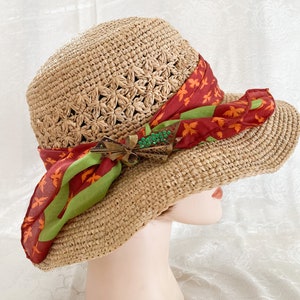 May include: A straw sun hat with a wide brim and a red and green patterned scarf tied around it. The hat is decorated with a gold and green brooch.