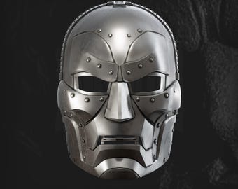 Puede incluir: Una máscara facial completa de metal plateado con un diseño segmentado y remaches visibles a lo largo de las cejas, los pómulos y la mandíbula. La máscara tiene aberturas rectangulares para los ojos y una pieza nasal central. El texto "STAVION STUDIOS" aparece debajo.