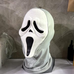 May include: A white Scream mask with black eye and mouth openings, set against a gray hood. The mask has a ghostly, elongated face shape, and the hood is a light gray color. The mask is a classic Halloween costume accessory.