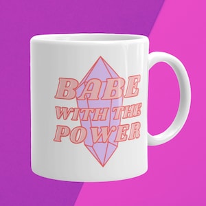 May include: White ceramic mug with a pink and purple design. The mug features a large purple crystal with a pink outline and the text "BABE WITH THE POWER" in pink.