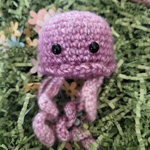 May include: A crocheted purple jellyfish with black eyes. The jellyfish is sitting on a bed of green and white shredded paper.