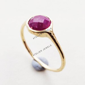 Op de afbeelding: Een gouden ring met een gefacetteerde, ovale robijn edelsteen. De ring heeft een eenvoudig ontwerp met de tekst "ATELIER JEWELS" op de ring.