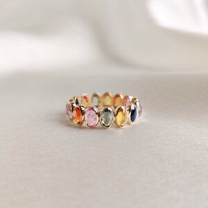 Puede incluir: Un anillo de banda de oro con una fila de piedras preciosas ovaladas en varios colores, incluyendo rosa, azul, verde, amarillo y naranja.