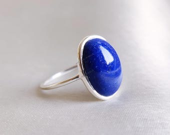 May include: A silver ring featuring a large, oval-shaped, deep blue gemstone. The gemstone is set in a silver bezel, and the ring has a simple, polished band. The ring is displayed against a soft, light background.