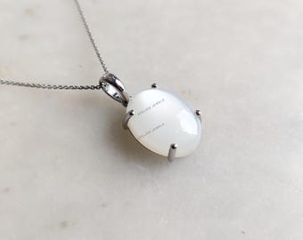 May include: A silver necklace with a large, oval, white gemstone pendant. The pendant is held in place by four silver prongs. The chain is delicate and the pendant has the text "ATELIER JEWELS".