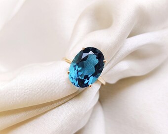 Può includere: Un anello d'oro con una grande gemma ovale blu-verde scuro. La gemma è incastonata con quattro griffe su una sottile fascia d'oro. L'anello è presentato su un tessuto bianco e setoso, creando un'estetica lussuosa.