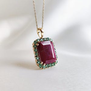 Op de afbeelding: Een gouden ketting met een rechthoekige, dieprode edelsteen hanger. De hanger is omgeven door kleine, groene edelstenen. De ketting hangt aan een fijne gouden ketting, een elegant sieraad.