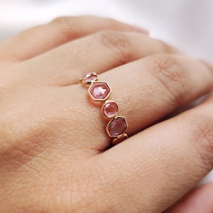Puede incluir: Un anillo de oro con una banda de piedras preciosas rosas. Las piedras preciosas son hexagonales y facetadas, y están engastadas en una banda de oro.