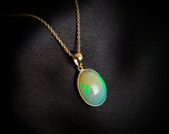 Puede incluir: Un collar con colgante de oro con una piedra preciosa de ópalo ovalada. El ópalo muestra una mezcla de tonos verdes, amarillos y azules. El collar tiene una fina cadena de oro y un engaste de oro alrededor del ópalo.