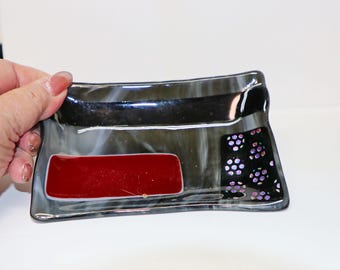 Könnte beinhalten: Eine dekorative Glasschale mit rechteckiger Form. Die Schale hat ein rotes Rechteck, einen schwarzen Bereich mit lila Punkten und ein grau-schwarzes Marmormuster. Die Schale wird von einer Hand gehalten.