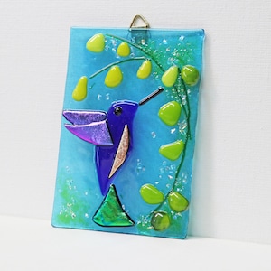 May include: A rectangular glass art piece featuring a blue hummingbird with purple wings and a green triangle tail. The background is a light blue with green and yellow accents, and a green vine with yellow and green teardrop shapes.