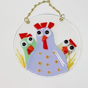 May include: A round glass ornament with a purple, green, and yellow chicken design. The chickens have large eyes and red combs. The ornament has a gold chain for hanging.