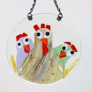 May include: A round glass ornament featuring three chickens. The chickens are made of stained glass and have large eyes, red combs, and orange beaks. The background is white with a straw-like texture.