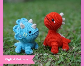 May include: Two crocheted dinosaur toys, one blue and one red, sitting on a bed of green grass. The blue dinosaur has three horns and a white spot pattern. The red dinosaur has a white spot on its head.