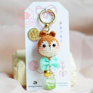 May include: A crocheted brown squirrel keychain with a light blue bow and a small bell. The keychain is attached to a gold key ring and a small round tag that says "Baby Squirrel". The keychain is hanging from a white card with Japanese text and the words "handmade crochet" and "amigurumi".