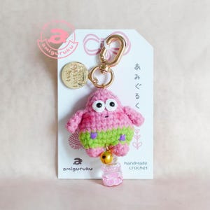 May include: A handmade crochet keychain featuring a pink amigurumi character with green shorts and purple accents. The keychain includes a gold-colored clasp, a small bell, and a clear charm with pink swirls. The tag reads "amiguruku" and "handmade crochet".