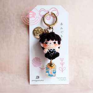 May include: A handmade crochet keychain featuring a small doll with black hair and a black outfit. The keychain includes a gold-colored clasp, a charm, and a small bell. The tag reads "amiguruku" and "handmade crochet".