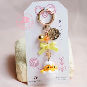 May include: A yellow crocheted chick peeking out of a white crocheted eggshell. The chick has black eyes and a small orange beak. The chick is attached to a gold key chain with a gold charm that says "Happy Every Day". The keychain also has a gold bell, a yellow bow, and a clear crystal bead.
