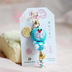 Puede incluir: Un llavero de crochet azul con un personaje de dibujos animados con la cara blanca y las mejillas rojas. El llavero tiene un dije de estrella dorada, un dije de campana dorada y un dije dorado con el texto "amigurumi" en japonés. El llavero está colgado de una tarjeta blanca con el texto "crochet hecho a mano" y "amigurumi" en japonés.