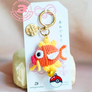 Puede incluir: Un llavero de pescado de ganchillo naranja y amarillo con una aleta blanca y una boca rosa. El llavero tiene un anillo dorado, un amuleto dorado con el texto "amigurumi" y un amuleto Poké Ball rojo y blanco. El llavero está colgado de una tarjeta blanca con el texto "100% hecho a mano amigurumi" y "あみぐるみ" en japonés.