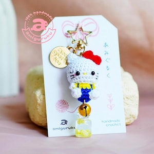 Puede incluir: Un llavero Hello Kitty de crochet blanco con un lazo rojo, un mono azul y una camisa amarilla. El llavero tiene un dije de estrella dorada, una campana dorada y un dije de botella transparente lleno de rodajas de limón amarillo. El llavero está hecho a mano y viene con una etiqueta que dice "100% amigurumi hecho a mano" y "crochet hecho a mano".