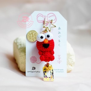 May include: Red crocheted Elmo keychain with a gold star charm, a gold bell, and a gold tag with Japanese text. The keychain is handmade and has a white yarn ball and a tag that says "amigurumi" in the bottom left corner.