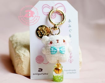 May include: White crocheted bear keychain with a blue and white bow, a gold bell, and a green and gold charm. The keychain is attached to a gold key ring and has a tag that says "amigurumi handmade crochet".