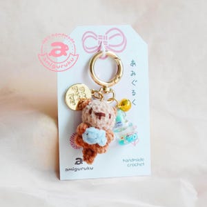 May include: A handmade crochet keychain featuring a small brown bear holding a blue object. The keychain has a gold-colored clasp and a small gold charm. The tag reads "amiguruku" and "handmade crochet".