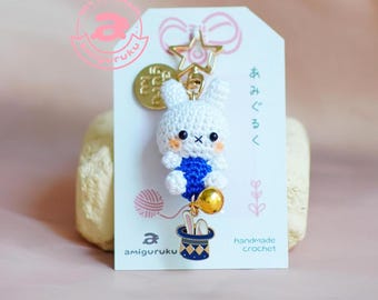 May include: White crocheted bunny rabbit wearing a blue outfit with a gold star charm and a gold bell hanging from a hat. The bunny is attached to a gold key chain. The card reads "handmade crochet" and "amigurumi".