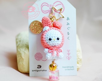 May include: A pink and white crocheted bunny keychain with a bell and a small glass bottle filled with pink and white beads. The keychain is attached to a gold star charm and a tag that reads "handmade crochet" and "amigurumi".